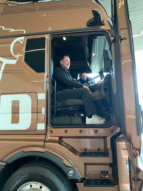 Zwei Männer sitzen lachend in der Fahrerkabine eines braunen Lastwagens mit offenem Türrahmen.